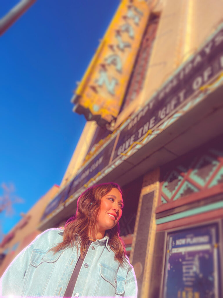 A pink glow engulfs Megan as she poses outside of the Mayan Theater's sign.